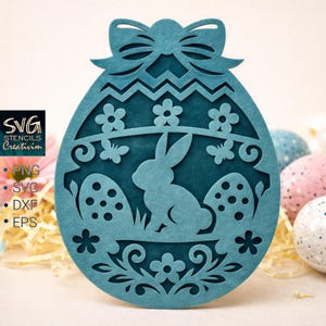May include: A teal Easter egg-shaped decoration with a bunny and floral cutouts. The decoration has a bow at the top and the text "SVG STENCILS Creativim" with file type options. Decorative Easter-themed craft.