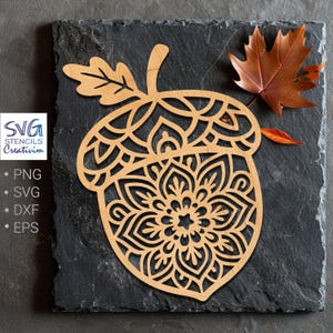 Peut inclure: Un gland en bois découpé au laser avec un motif mandala détaillé, présenté sur une surface en ardoise gris foncé. Le gland est accompagné d'une feuille d'automne brune. L'image comprend également le texte "SVG Stencils Creativim".