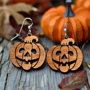 Peut inclure: Boucles d'oreilles en bois en forme de citrouille avec crochets argent&eacute;s. Chaque boucle d'oreille est sculpt&eacute;e en forme de lanterne d'Halloween souriante. Les boucles d'oreilles sont pr&eacute;sent&eacute;es sur une surface en bois avec des citrouilles et des feuilles d'automne.