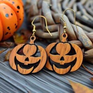 Peut inclure: Boucles d'oreilles en bois en forme de citrouille avec des crochets dor&eacute;s. Les boucles d'oreilles sont sculpt&eacute;es avec un visage de lanterne et sont orange et noir. Une citrouille d&eacute;corative orange et des feuilles d'automne sont en arri&egrave;re-plan.