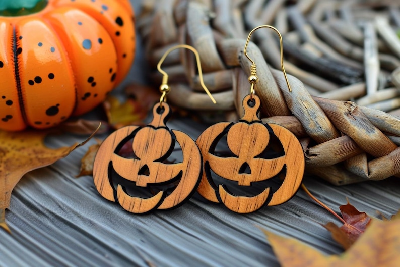 Peut inclure: Boucles d'oreilles en bois en forme de citrouille avec des crochets dor&eacute;s. Les boucles d'oreilles sont sculpt&eacute;es avec un visage de lanterne et sont orange et noir. Une citrouille d&eacute;corative orange et des feuilles d'automne sont en arri&egrave;re-plan.