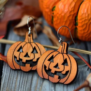 Peut inclure: Deux boucles d'oreilles en bois en forme de citrouille avec des crochets argent&eacute;s. Les boucles d'oreilles sont orange avec des visages de jack-o'-lantern sculpt&eacute;s. Le fond comprend des feuilles d'automne et une citrouille sculpt&eacute;e, sur le th&egrave;me d'Halloween.
