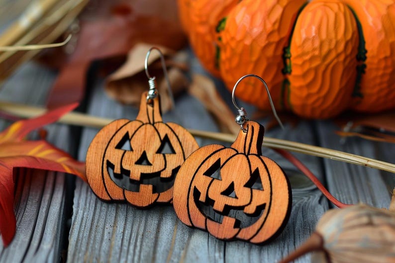 Peut inclure: Deux boucles d'oreilles en bois en forme de citrouille avec des crochets argent&eacute;s. Les boucles d'oreilles sont orange avec des visages de jack-o'-lantern sculpt&eacute;s. Le fond comprend des feuilles d'automne et une citrouille sculpt&eacute;e, sur le th&egrave;me d'Halloween.