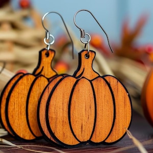 Peut inclure: Boucles d'oreilles en bois en forme de citrouille avec des crochets argent&eacute;s. Les boucles d'oreilles sont orange avec des contours marron fonc&eacute;. Une petite citrouille orange est &agrave; l'arri&egrave;re-plan, avec de l'herbe s&eacute;ch&eacute;e et des baies rouges.