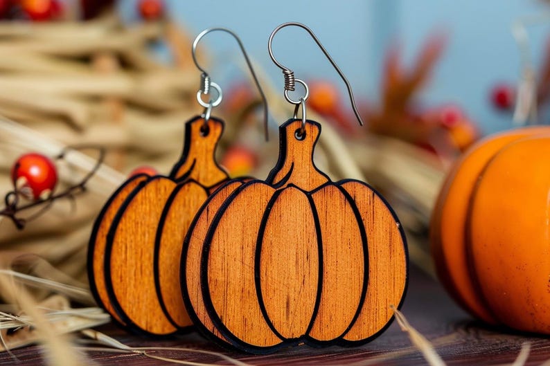 Peut inclure: Boucles d'oreilles en bois en forme de citrouille avec des crochets argent&eacute;s. Les boucles d'oreilles sont orange avec des contours marron fonc&eacute;. Une petite citrouille orange est &agrave; l'arri&egrave;re-plan, avec de l'herbe s&eacute;ch&eacute;e et des baies rouges.