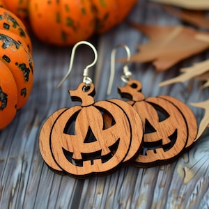 Peut inclure: Boucles d'oreilles en bois en forme de citrouilles souriantes avec des crochets en argent. L'arri&egrave;re-plan pr&eacute;sente des citrouilles oranges et des feuilles d'automne, parfaites pour Halloween.