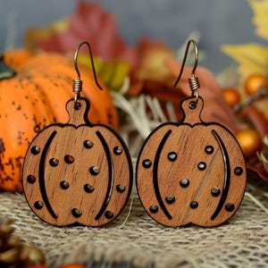 Peut inclure: Deux boucles d'oreilles en bois en forme de citrouille avec des crochets d'oreille cuivr&eacute;s. Les boucles d'oreilles sont sculpt&eacute;es en forme de citrouille, avec des lignes courbes et de petites d&eacute;coupes circulaires. L'arri&egrave;re-plan comprend une citrouille orange, des feuilles d'automne et des &eacute;l&eacute;ments d&eacute;coratifs.