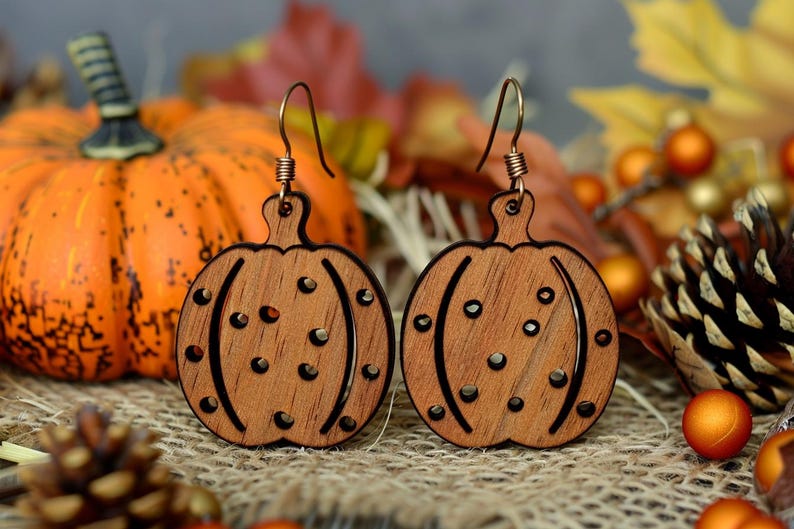 Peut inclure: Deux boucles d'oreilles en bois en forme de citrouille avec des crochets d'oreille cuivr&eacute;s. Les boucles d'oreilles sont sculpt&eacute;es en forme de citrouille, avec des lignes courbes et de petites d&eacute;coupes circulaires. L'arri&egrave;re-plan comprend une citrouille orange, des feuilles d'automne et des &eacute;l&eacute;ments d&eacute;coratifs.