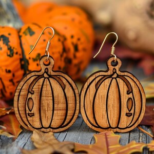 Peut inclure: Une paire de boucles d'oreilles en bois en forme de citrouille avec des crochets dor&eacute;s. Les boucles d'oreilles sont marron clair avec des d&eacute;tails grav&eacute;s. Le fond comprend des citrouilles oranges et des feuilles d'automne.