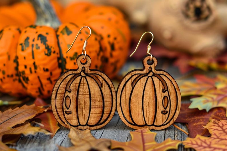 Peut inclure: Une paire de boucles d'oreilles en bois en forme de citrouille avec des crochets dor&eacute;s. Les boucles d'oreilles sont marron clair avec des d&eacute;tails grav&eacute;s. Le fond comprend des citrouilles oranges et des feuilles d'automne.