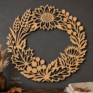 May include: A circular laser-cut wooden wall art piece featuring a detailed botanical wreath design. The cutout displays a sunflower, wheat stalks, thistle, and clusters of berries arranged in a ring. The text Digital Cut Files is visible.