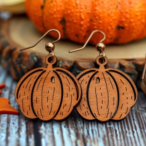 Peut inclure: Boucles d'oreilles en bois en forme de citrouille avec des crochets de couleur cuivre. Les boucles d'oreilles sont de couleur marron chaud et pr&eacute;sentent des lignes grav&eacute;es pour ressembler &agrave; une citrouille. Le fond comprend une citrouille orange et des feuilles d'automne.