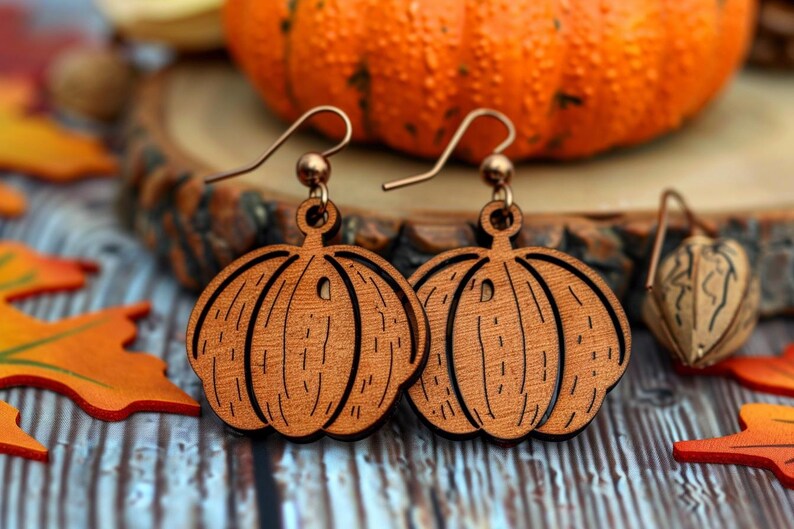 Peut inclure: Boucles d'oreilles en bois en forme de citrouille avec des crochets de couleur cuivre. Les boucles d'oreilles sont de couleur marron chaud et pr&eacute;sentent des lignes grav&eacute;es pour ressembler &agrave; une citrouille. Le fond comprend une citrouille orange et des feuilles d'automne.