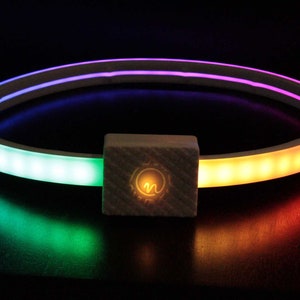 May include: A white circular LED light with a small white square control box in the center. The light is illuminated in a rainbow pattern of purple, blue, green, yellow, orange, and red.
