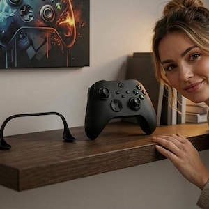 May include: A black Xbox controller rests on a dark wood shelf. A black curved object sits to the left of the controller. A framed image of a video game controller hangs on the wall above the shelf.