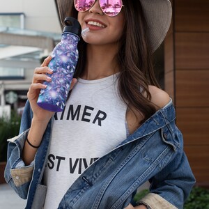 May include: A woman wearing a straw hat with pink mirrored sunglasses is holding a purple and blue water bottle with a straw. She is wearing a white tank top with the text "SUMMER" and "ST VIV" printed on it. She is also wearing a blue denim jacket.