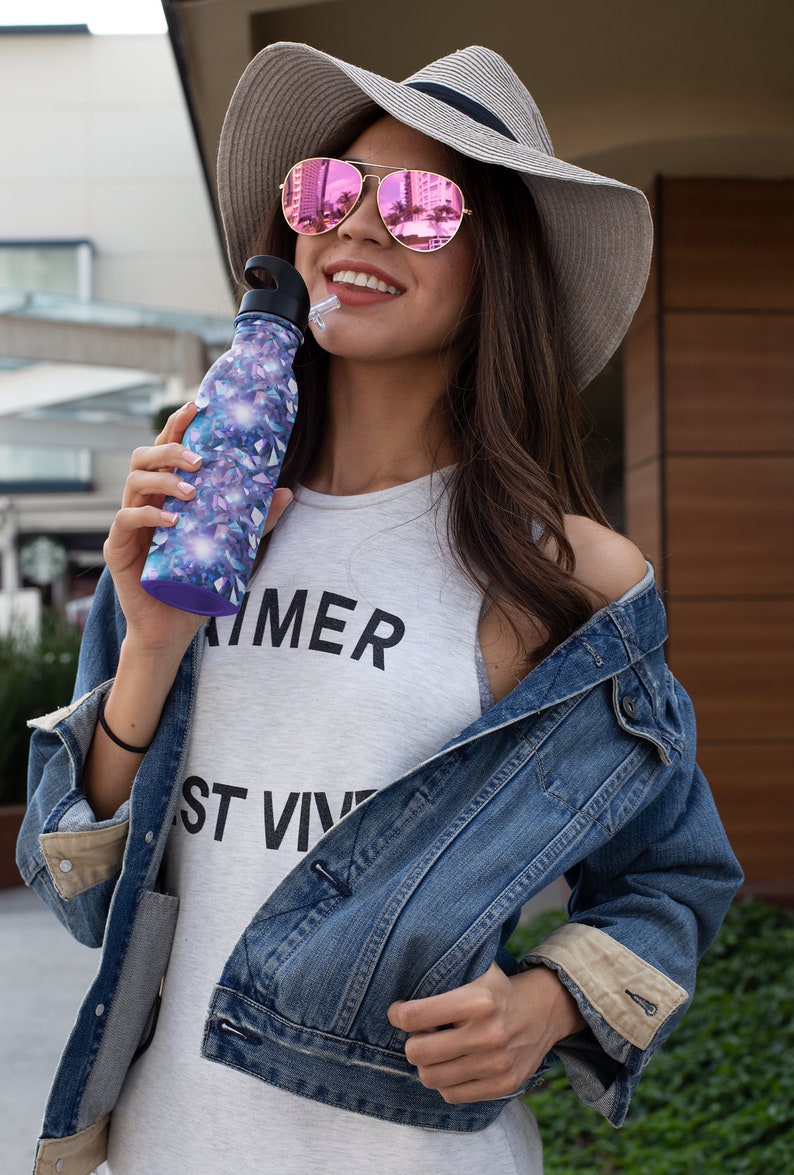 May include: A woman wearing a straw hat with pink mirrored sunglasses is holding a purple and blue water bottle with a straw. She is wearing a white tank top with the text "SUMMER" and "ST VIV" printed on it. She is also wearing a blue denim jacket.