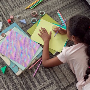 May include: A young person is lying on a carpet and drawing on a yellow piece of paper with a purple pencil. The paper is on top of a green folder. There are several colorful pencils, a book, and other school supplies scattered around the person.