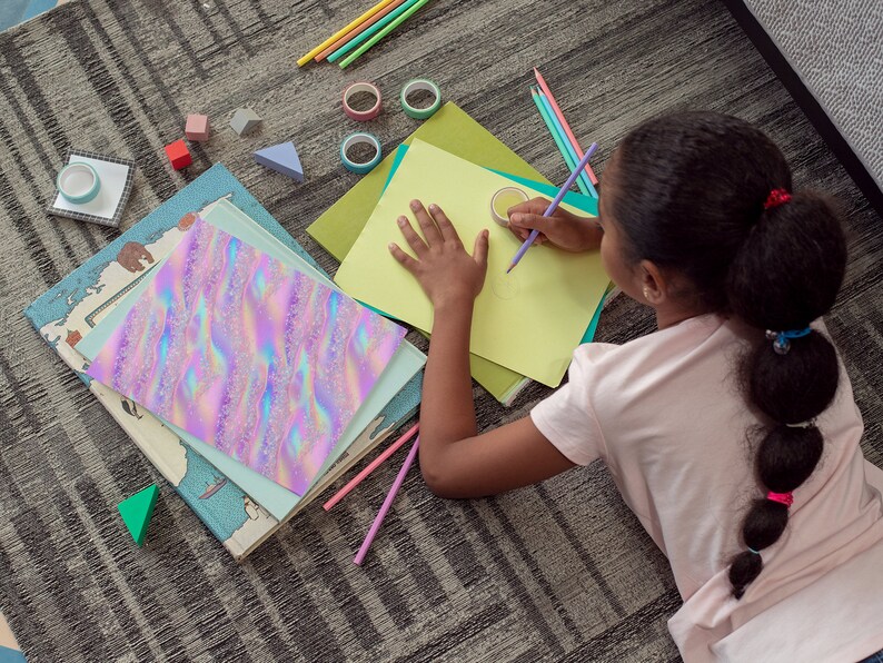 May include: A young person is lying on a carpet and drawing on a yellow piece of paper with a purple pencil. The paper is on top of a green folder. There are several colorful pencils, a book, and other school supplies scattered around the person.
