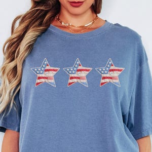 Puede incluir: Una camiseta azul con tres estrellas bordadas. Cada estrella presenta un diseño de patchwork con patrones rojos, blancos y azules, evocando la bandera estadounidense. La camiseta tiene cuello redondo y mangas cortas.