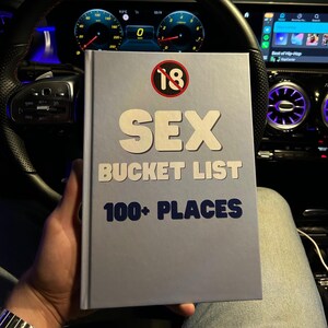 May include: A light gray book titled "SEX BUCKET LIST" with the text "100+ PLACES" in blue. The cover also features a red circle with the number "18" inside and crossed out. The book is held in front of a car's dashboard.