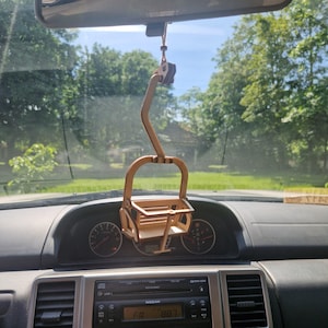 Pode incluir: Um ambientador de carro em forma de teleférico de madeira pendurado no espelho retrovisor. O teleférico é feito de madeira clara e tem um design detalhado. O fundo mostra árvores e um céu azul.