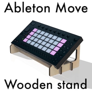 Op de afbeelding: Zwart elektronisch muziekproductieapparaat op een houten standaard. Het apparaat heeft een raster van verlichte pads in wit en roze. De standaard is gemaakt van lichtgekleurd hout, met de tekst "Ableton Move" en "Wooden stand".