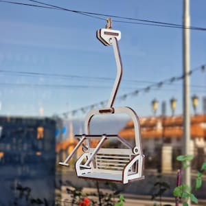 May include: A wooden ski lift chair, painted white with brown trim, hangs from a wire. The chair has a seat, back, and safety bar. The background is a blurred view of a building and sky.
