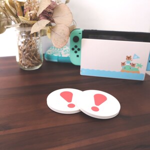 May include: Two white round coasters with a red exclamation point on each. The coasters are on a wooden table next to a blue Nintendo Switch console with a Animal Crossing themed case.