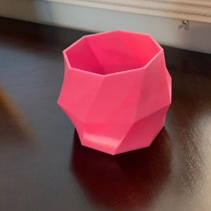 Op de afbeelding: Een roze, geometrisch gevormde plantenbak met een facetontwerp. De plantenbak is gemaakt van een solide materiaal en heeft een brede opening. De plantenbak staat op een donkere ondergrond.