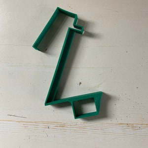 Könnte beinhalten: Ein grüner Ausstecher aus Kunststoff in Form der Zahl eins. Der Ausstecher ist einteilig mit flachem Boden und rechteckiger Oberseite. Er liegt auf einer weißen Holzoberfläche.