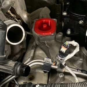 May include: An engine bay with various components. A bright red cap sits atop a metal component. Silver tubes and black hoses are visible, along with electrical wiring and connectors. The overall scene suggests automotive repair or maintenance.