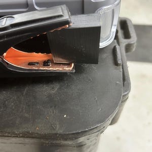 May include: A black plastic clamp with copper-colored metal teeth gripping a small, rectangular black block. The clamp is positioned against a gray, textured plastic case with a translucent, ribbed side panel and a black handle.