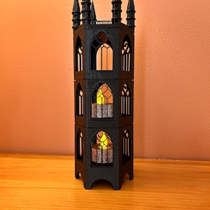 Könnte beinhalten: Ein schwarzer, dreistöckiger gotischer Turm-Kerzenhalter mit spitzen Türmchen. Der Kerzenhalter hat gewölbte Fenster und enthält ein flackerndes, warm getöntes Licht. Die Struktur besteht aus dunklem Material und steht auf einer Holzoberfläche.