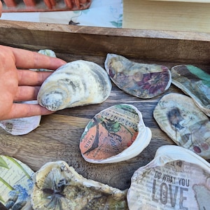 以下が含まれることがあります： ヴィンテージ風の紙デコパージュが施された、装飾されたカキ殻のコレクション。 殻は木製のトレイに配置されています。 殻の1つには「PARIS」という文字が印刷されています。 他の殻には「DO WHAT YOU LOVE ART」という文字が印刷されています。