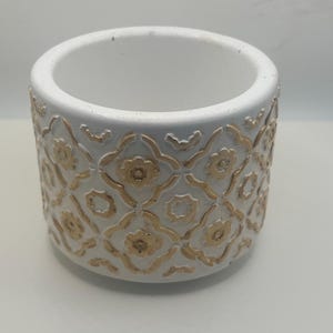 May include: A white ceramic planter with a gold floral pattern. The cylindrical pot has a wide opening and a textured surface. The gold design is embossed on the white background, creating a decorative effect. Ideal for small plants or succulents.