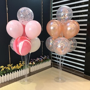 May include: Two balloon bouquets, one with pink and white balloons, the other with peach and clear confetti balloons. The balloons are arranged on clear stands, suitable for party decorations. The background includes a white picket fence and a black wall.