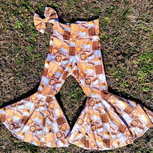 Op de afbeelding: Een roze en bruine broek met wijde pijpen met een s'mores-patroon. De broek heeft een wijde pijp en een hoge taille. Het patroon bevat cartoonafbeeldingen van s'mores met marshmallows, chocolade en graham crackers.