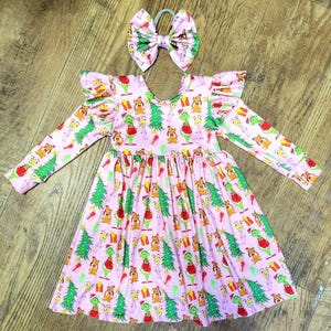 Könnte beinhalten: Ein rosa Kleid mit langen Ärmeln, Rüschen am Ausschnitt und an den Ärmeln. Das Kleid hat ein sich wiederholendes Muster aus grünen Weihnachtsbäumen, roten Geschenken und einem grünen Wesen, das eine rote Weihnachtsmütze trägt. Ein rosa Schleife mit einer weißen Mitte ist enthalten.
