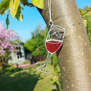 Puede incluir: Un adorno de vidrio de vino de vidrio coloreado con un relleno de vino tinto que cuelga de una rama de árbol. El vidrio es plateado y el vino es rojo oscuro.