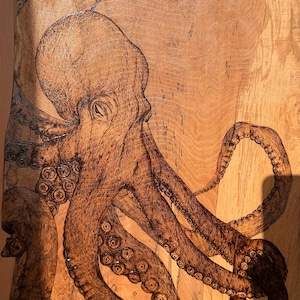 May include: A wood-burned artwork of an octopus, featuring detailed lines and shading. The octopus is rendered in brown tones, with visible suckers on its tentacles. The artwork is on a light brown wooden surface, showing the wood grain.