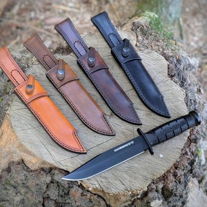Puede incluir: Un cuchillo negro con el texto "LEATHERNECK-SF" y un mango negro se muestra con cuatro fundas de cuero en tonos marrones. Las fundas están dispuestas sobre un tocón de árbol.