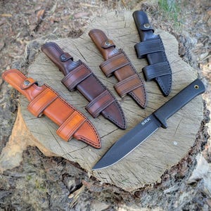 Op de afbeelding: Een assortiment messen en scheden op een boomstronk. De messen hebben zwarte bladen en zwarte handvatten. De scheden zijn van leer in verschillende bruin- en zwarttinten, met riemen en knoopsluitingen.