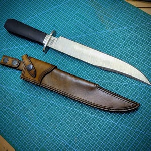 May include: A large, stainless steel knife with a black handle and a brown leather sheath. The knife has a wide blade and a silver bolster. The sheath has a belt loop with two snap closures. The knife and sheath are on a green cutting mat.