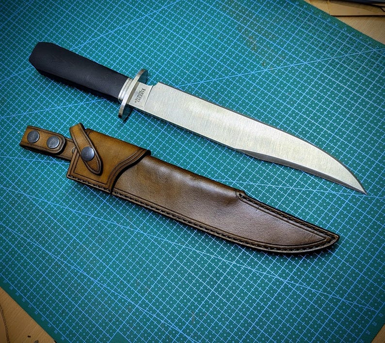 May include: A large, stainless steel knife with a black handle and a brown leather sheath. The knife has a wide blade and a silver bolster. The sheath has a belt loop with two snap closures. The knife and sheath are on a green cutting mat.