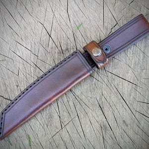 May include: A brown leather knife sheath with a matching belt loop and snap closure. The sheath is stitched along the edges and is designed to hold a knife. The leather has a natural, worn appearance.