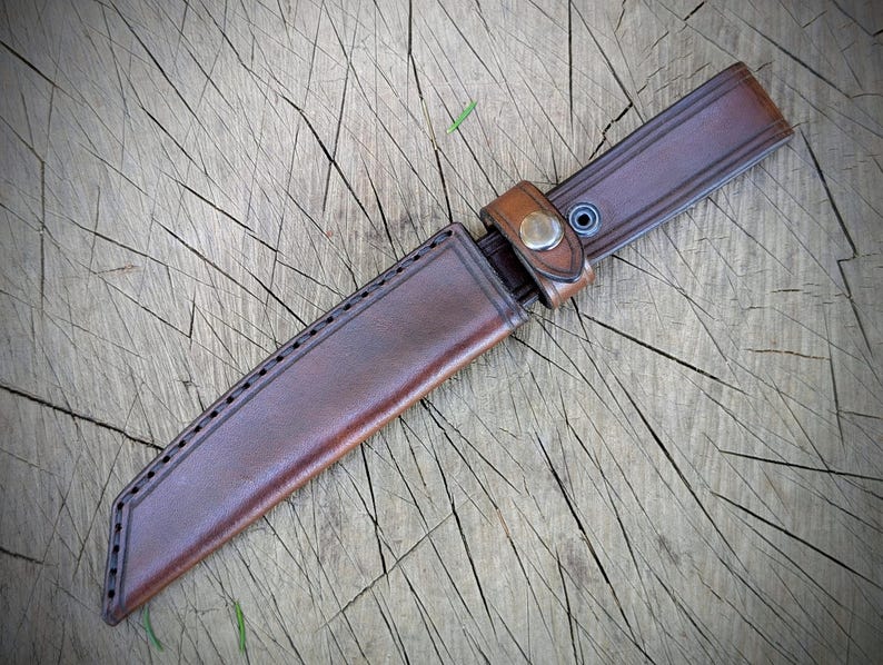 May include: A brown leather knife sheath with a matching belt loop and snap closure. The sheath is stitched along the edges and is designed to hold a knife. The leather has a natural, worn appearance.