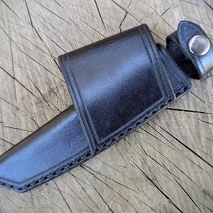 May include: A black leather knife sheath with a snap closure. The sheath is designed to hold a knife and has a belt loop. The leather is stitched along the edges.