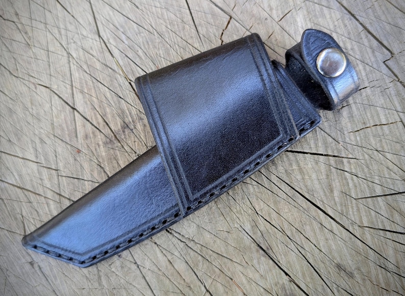 May include: A black leather knife sheath with a snap closure. The sheath is designed to hold a knife and has a belt loop. The leather is stitched along the edges.