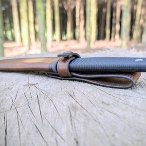 May include: A black knife with a textured handle is secured in a brown leather sheath with a matching belt loop. The knife and sheath are resting on a weathered wooden surface, with a blurred forest background.